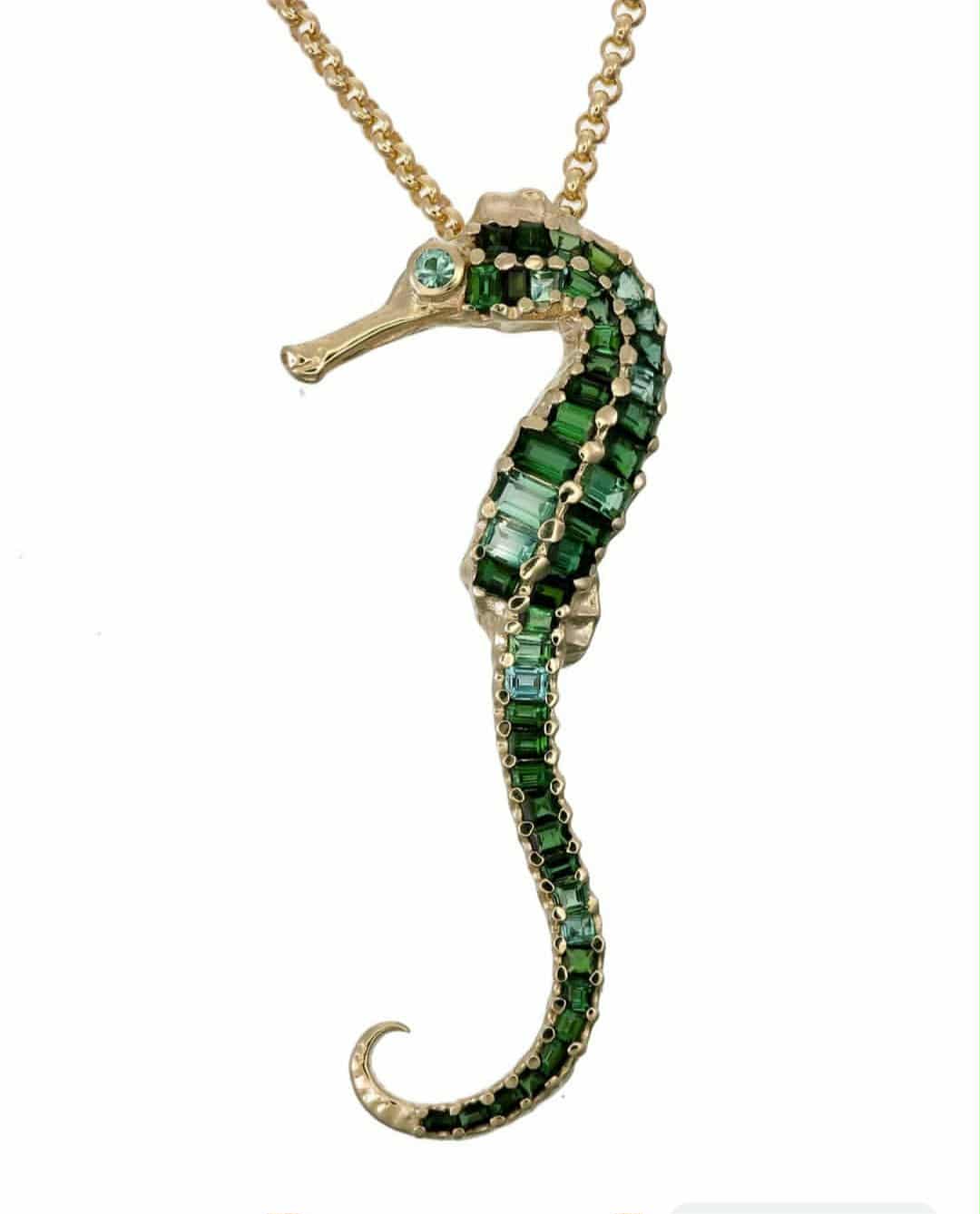Jewelry pendant shaped like a seahorse with green gemstones on a gold chain against a white background